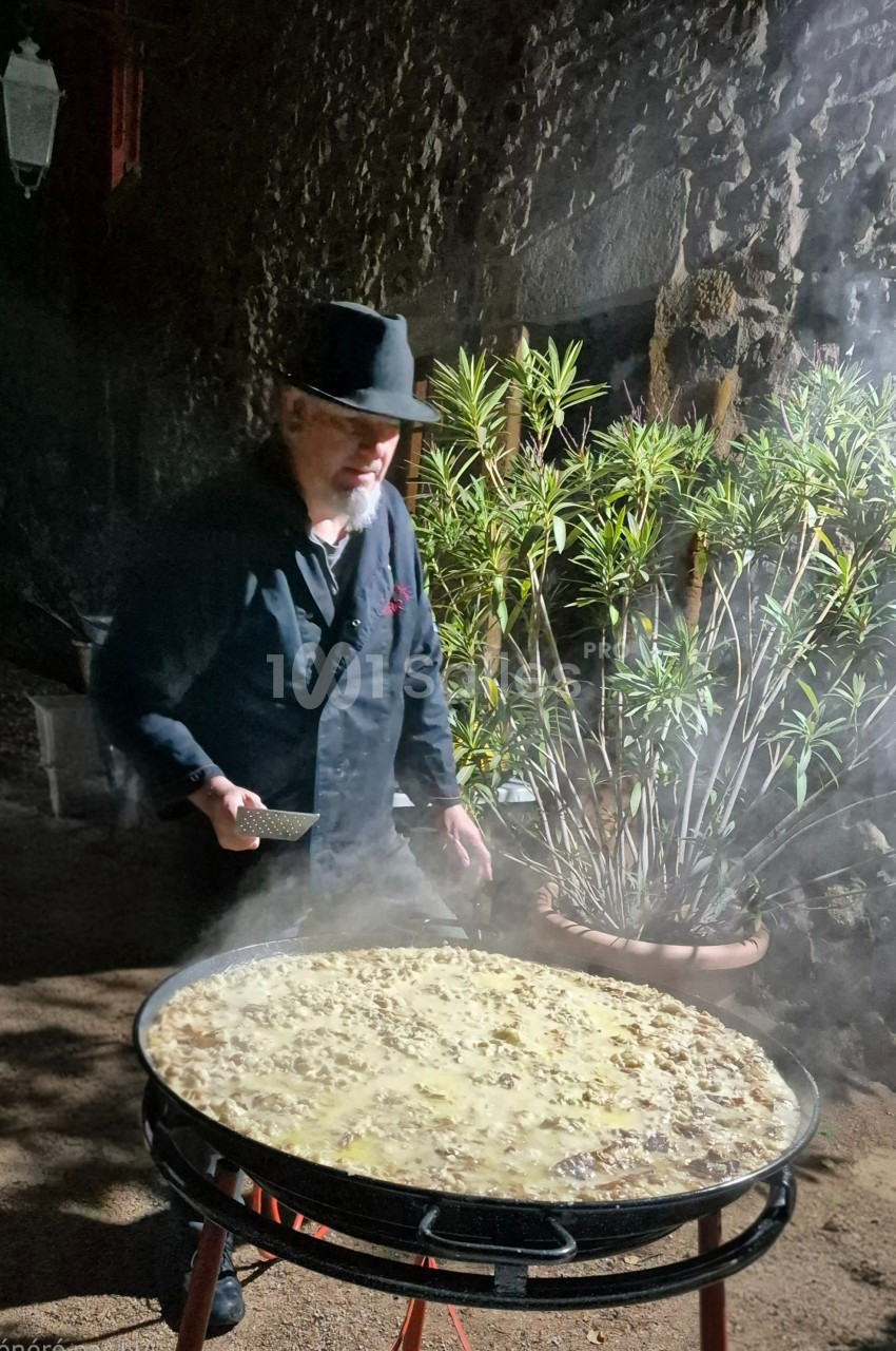 Un homme cuisine une grande paella fumante en extérieur, près d'un mur en pierre et de plantes en pot.