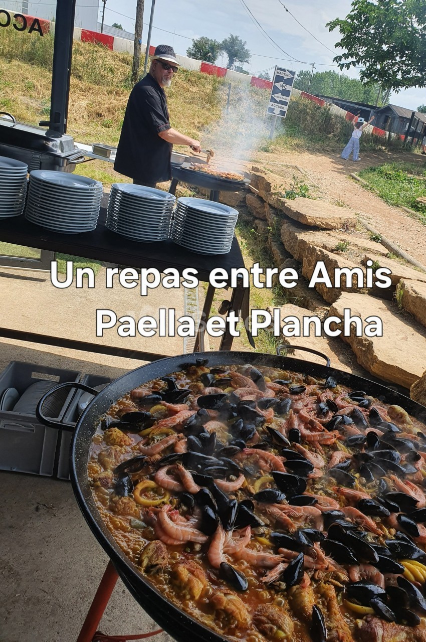 Un cuisinier prépare un repas en plein air avec une grande paella garnie de fruits de mer au premier plan.