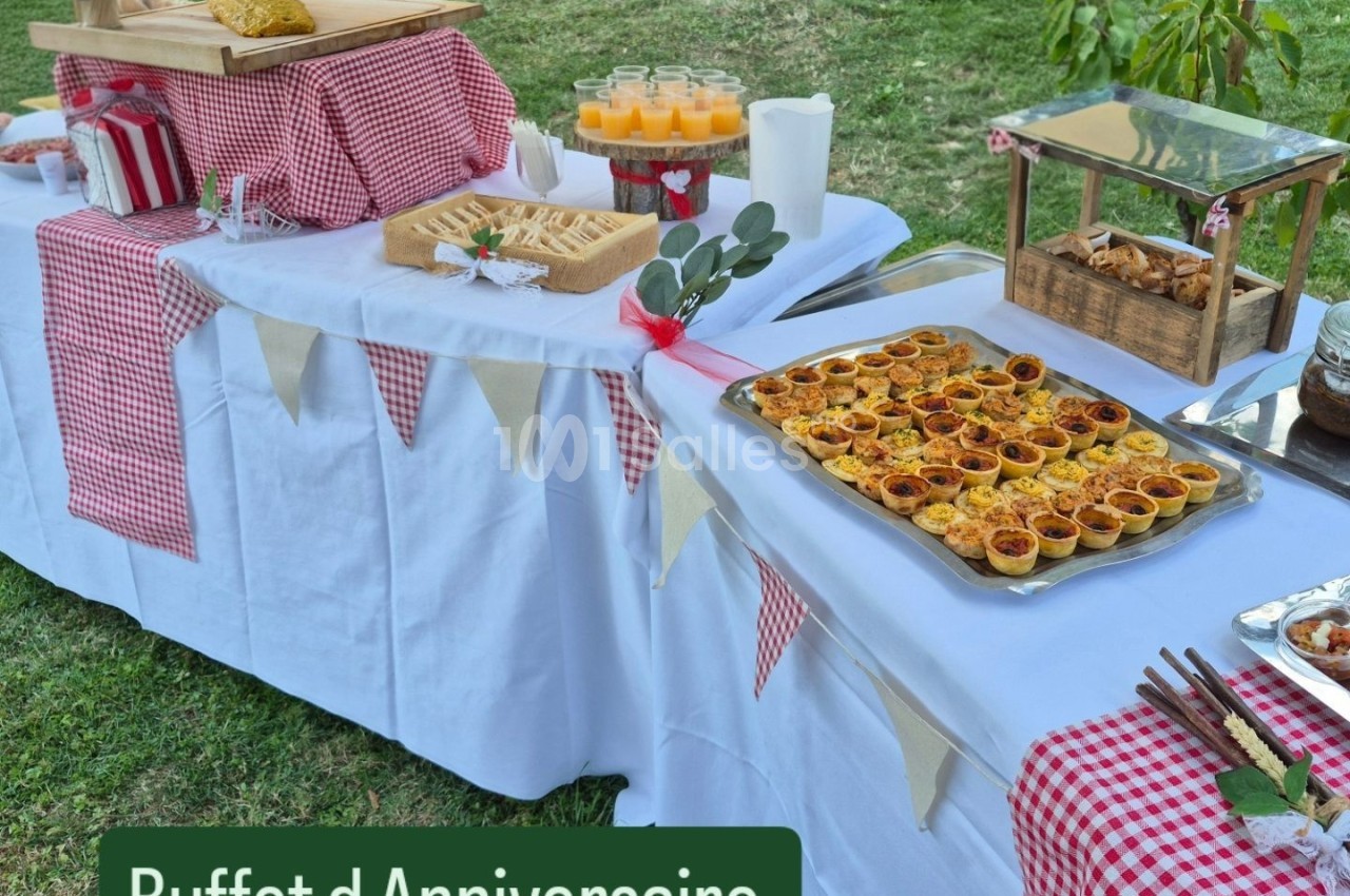 Buffet d'anniversaire en extérieur avec nappes blanches, plats variés, boissons et décor champêtre.