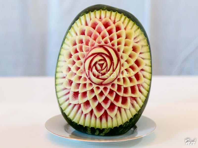Pastèque sculptée avec un motif floral complexe, posée sur une assiette blanche devant un fond clair.