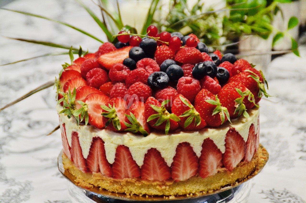 Gâteau décoré de fraises, framboises, myrtilles et groseilles sur une table avec un fond de plantes vertes.