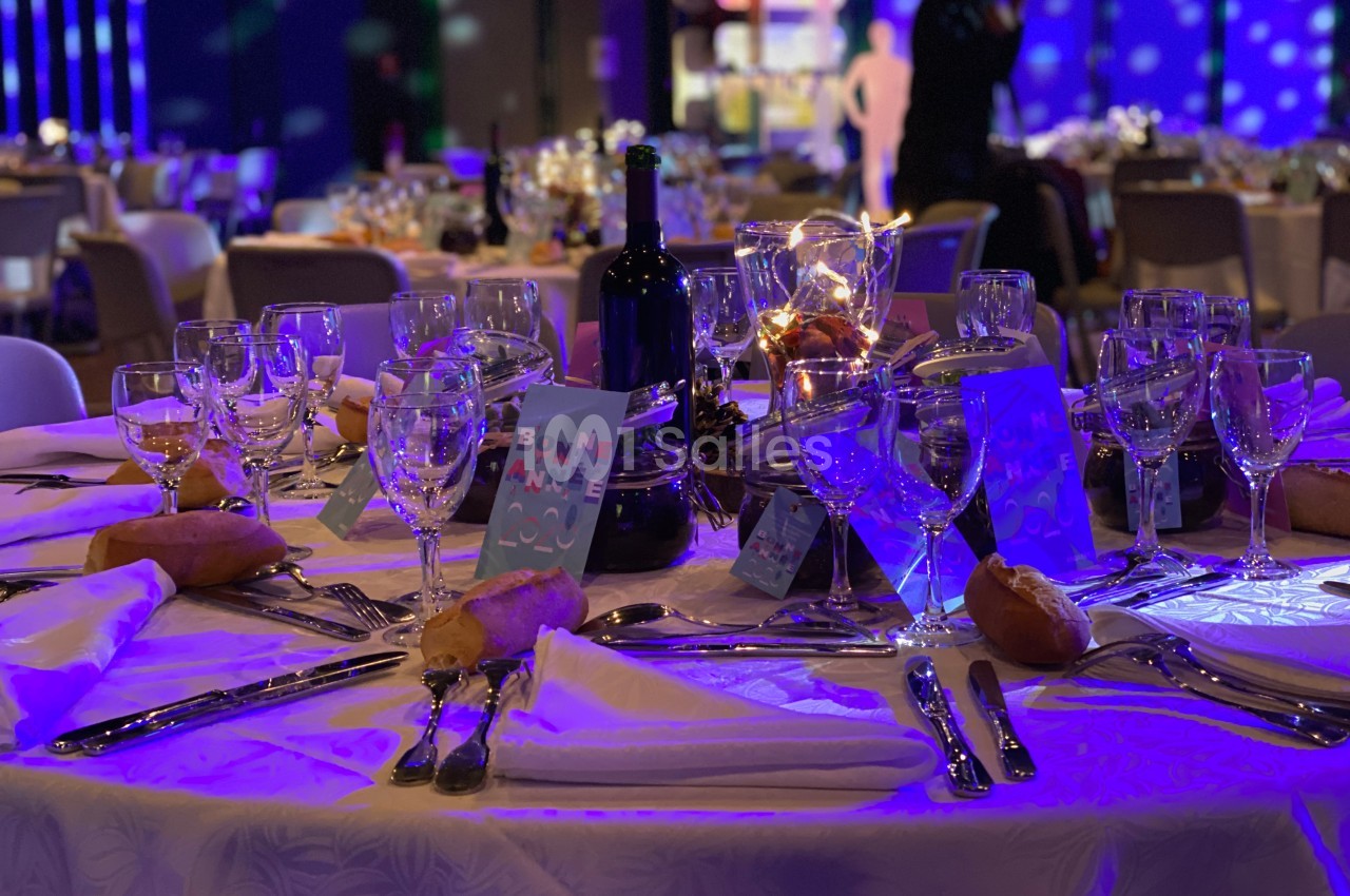 Table dressée pour un dîner festif avec nappes blanches, verres, couverts, bougies et décorations lumineuses.