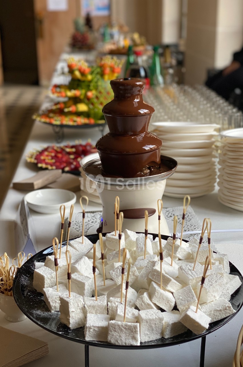 Fontaine de chocolat entourée de brochettes de guimauves et d'autres desserts disposés sur une table de réception.