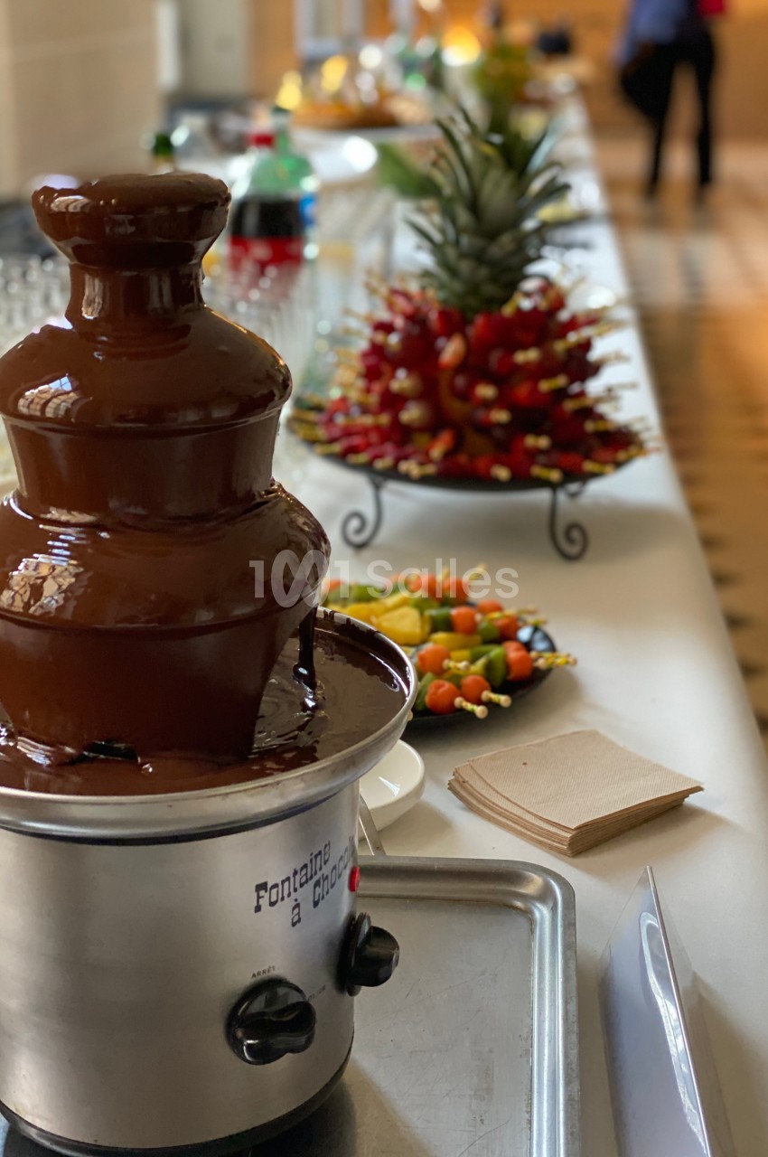 Fontaine à chocolat sur une table de buffet avec des fruits frais et des brochettes en arrière-plan.