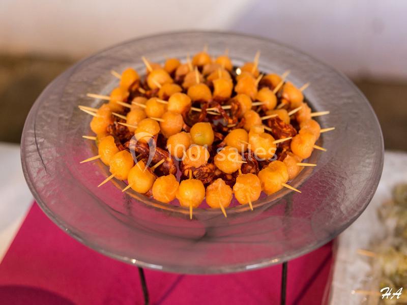 Assiette en verre contenant des brochettes de billes de melon et morceaux de charcuterie, présentée sur une nappe rouge.