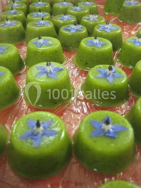 Petits desserts verts décorés de fleurs bleues, disposés en rangées sur un support transparent.