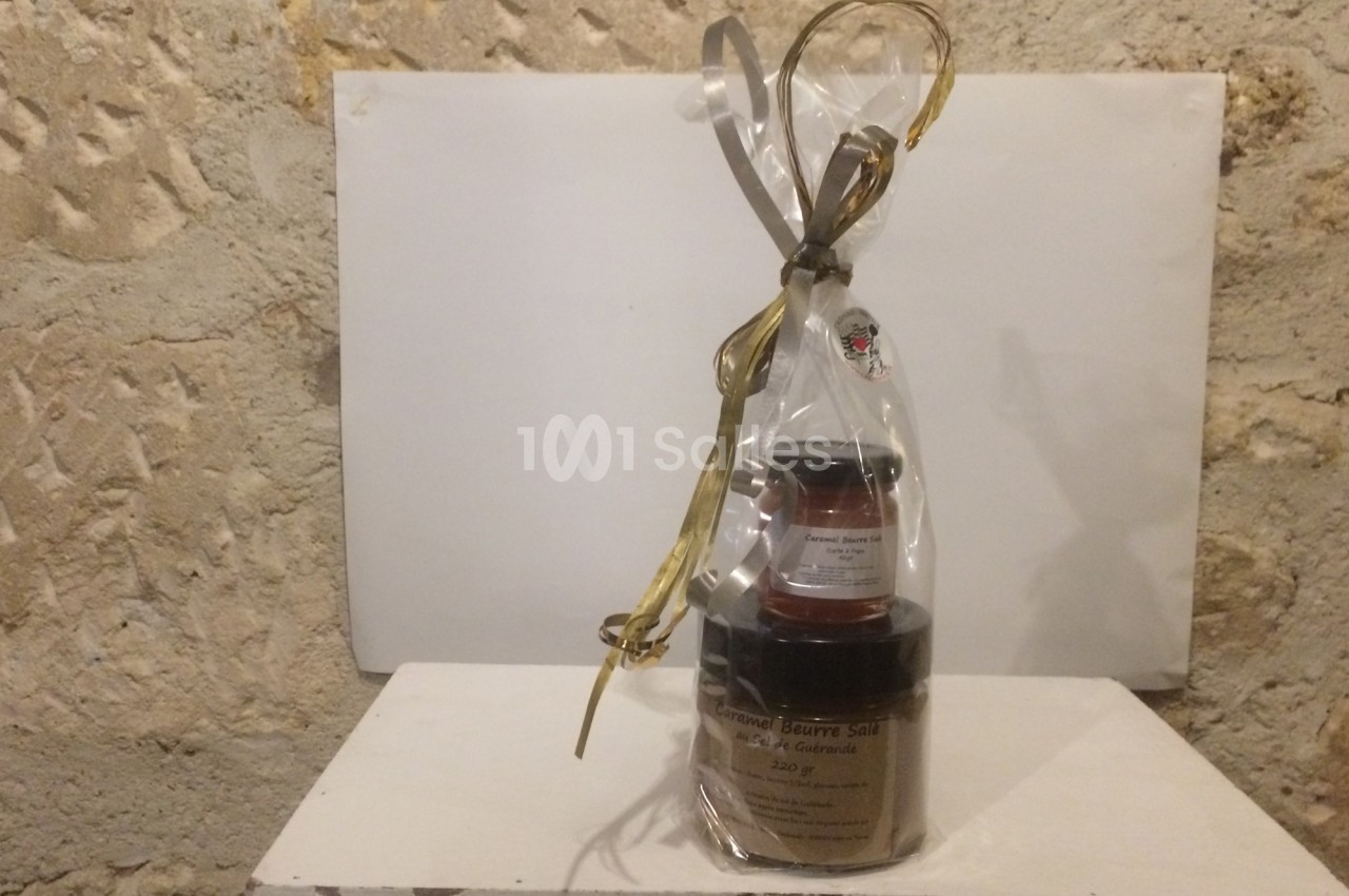 Ensemble de pots de confiture et beurre salé emballés dans un sachet transparent avec ruban doré, posé sur une table blanche.