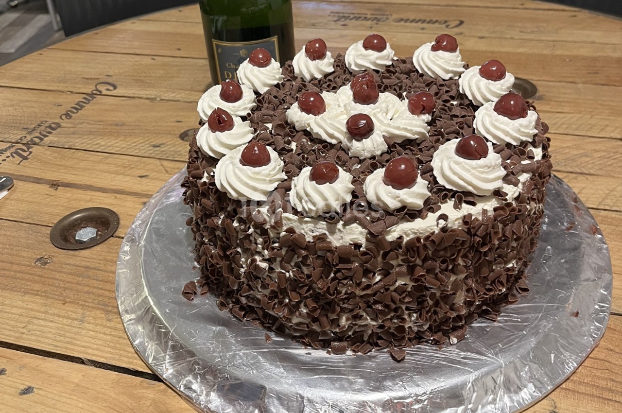 Gâteau forêt-noire décoré de crème chantilly, cerises confites et copeaux de chocolat, posé sur une table en bois.