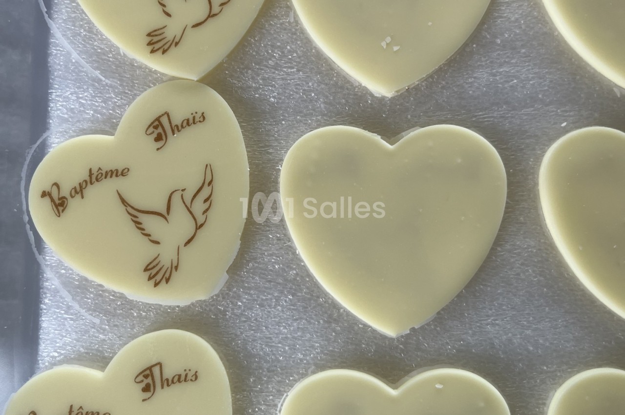 Cœurs en chocolat blanc, certains gravés d'une colombe et du texte ’Baptême Thaïs’, posés sur une surface argentée.