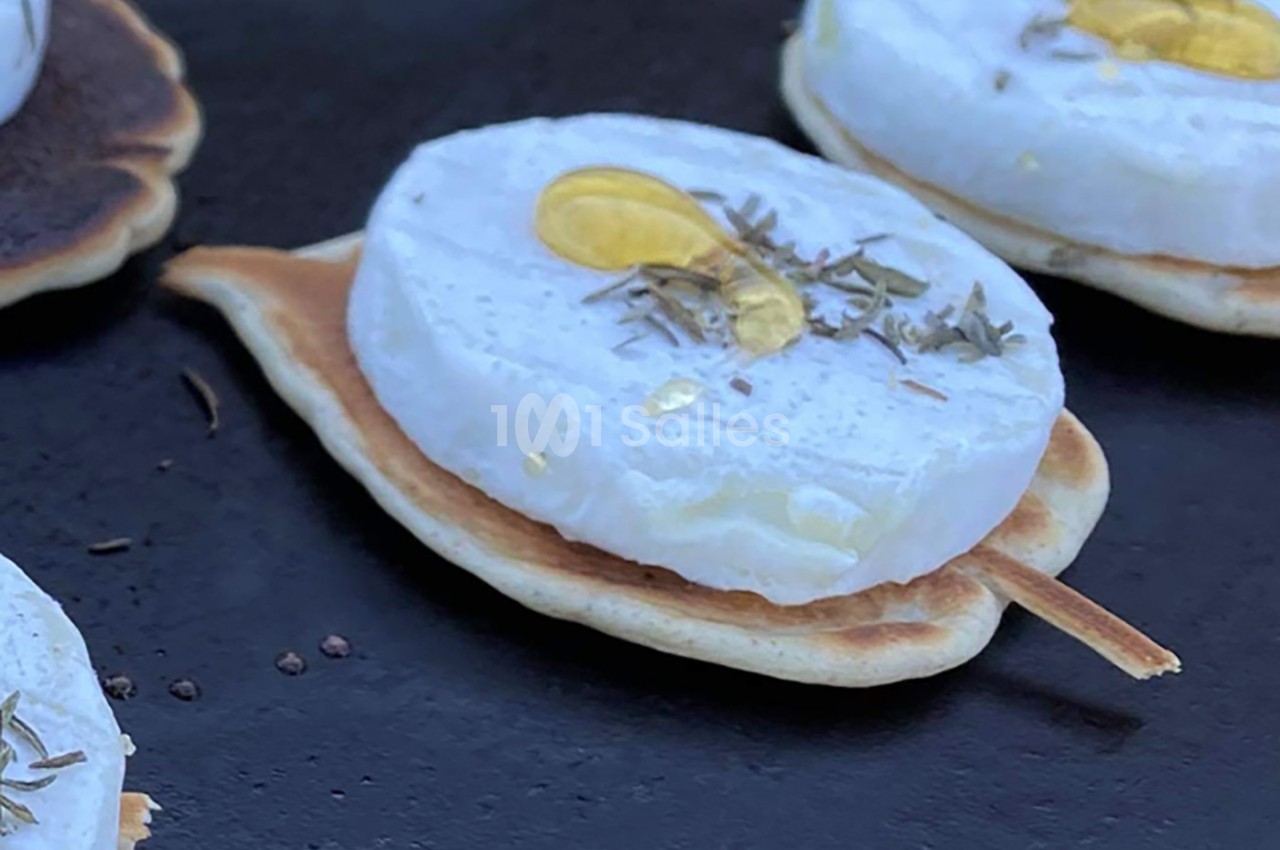 Tranche de fromage fondant sur un mini-blini, garnie de miel et d'herbes, présentée sur une surface noire.