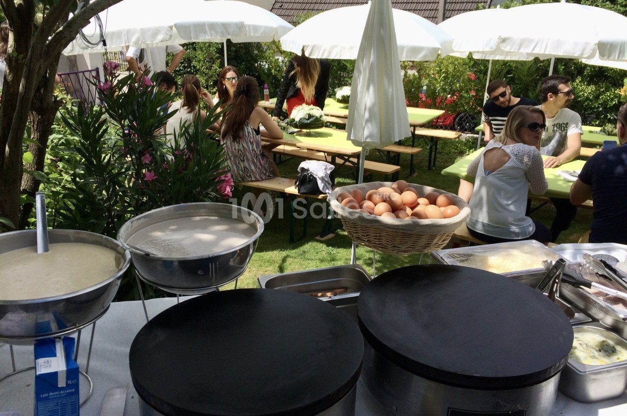 Stand de crêpes en extérieur avec des tables de convives sous des parasols dans un jardin verdoyant.