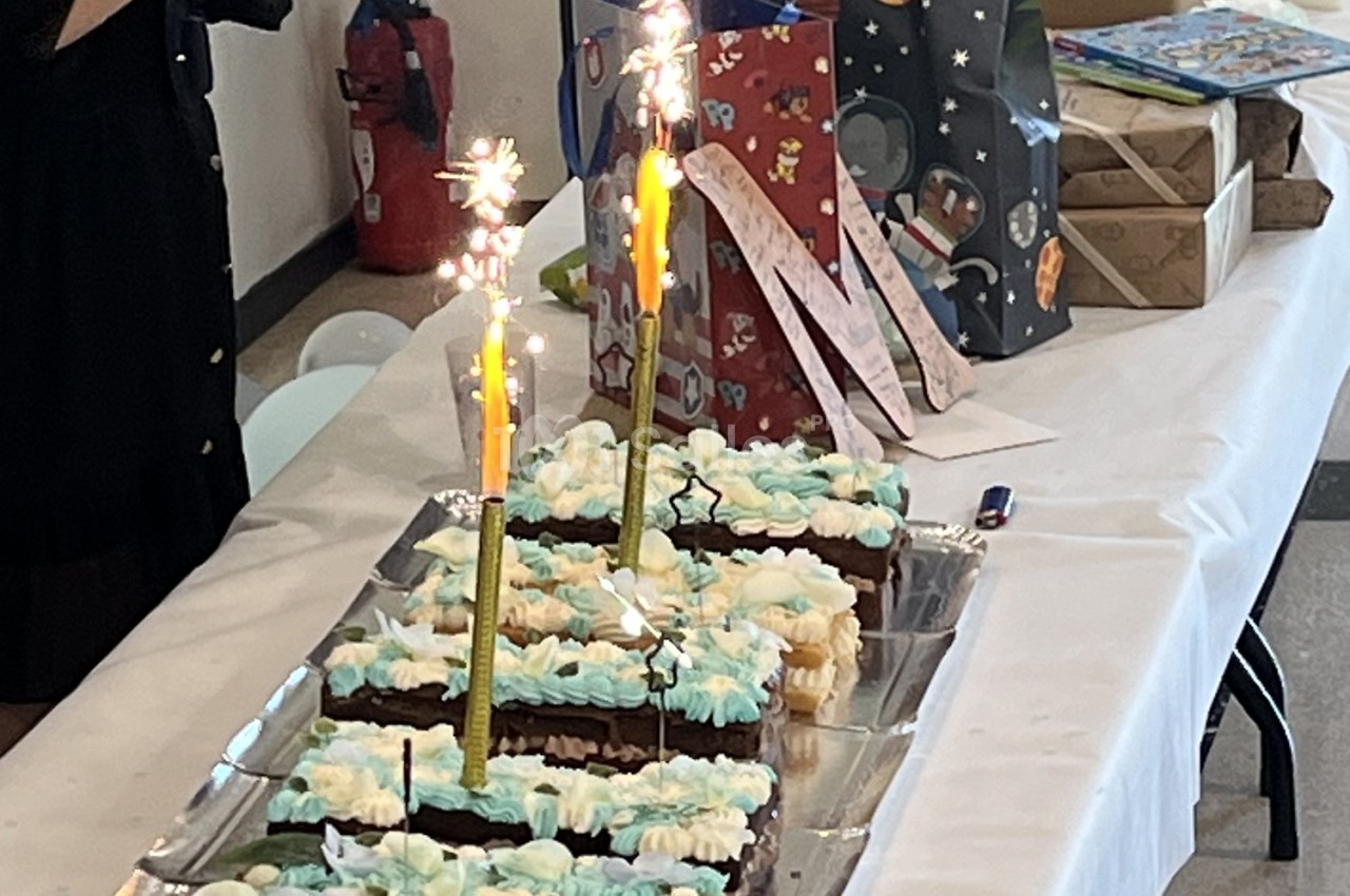 Gâteau rectangulaire décoré de crème bleue et blanche avec bougies étincelantes, entouré de cadeaux sur une table.
