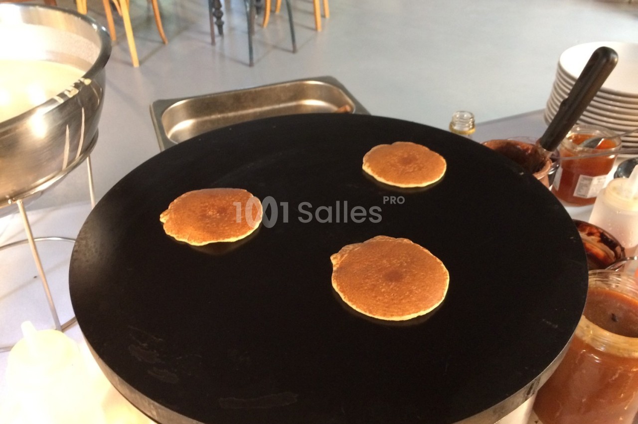 Trois pancakes cuisent sur une grande plaque chauffante noire dans un espace de cuisine.