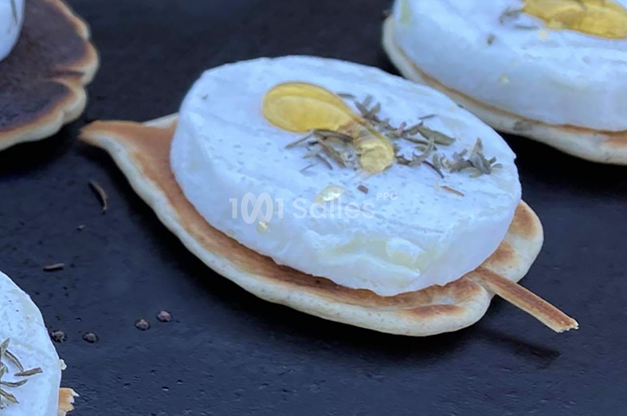 Tranche de fromage fondant sur un mini-blini, garnie de miel et d'herbes, présentée sur une surface noire.
