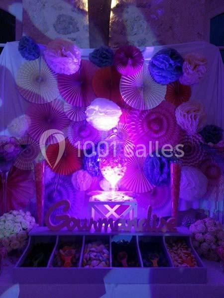 Mur décoratif avec éventails colorés, fleurs en papier, éclairage violet et présentoir de bonbons marqué ’Gourmandise’.