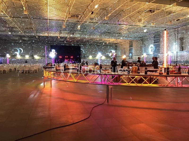 Salle de réception spacieuse avec éclairages colorés, tables dressées et un long bar illuminé au centre.