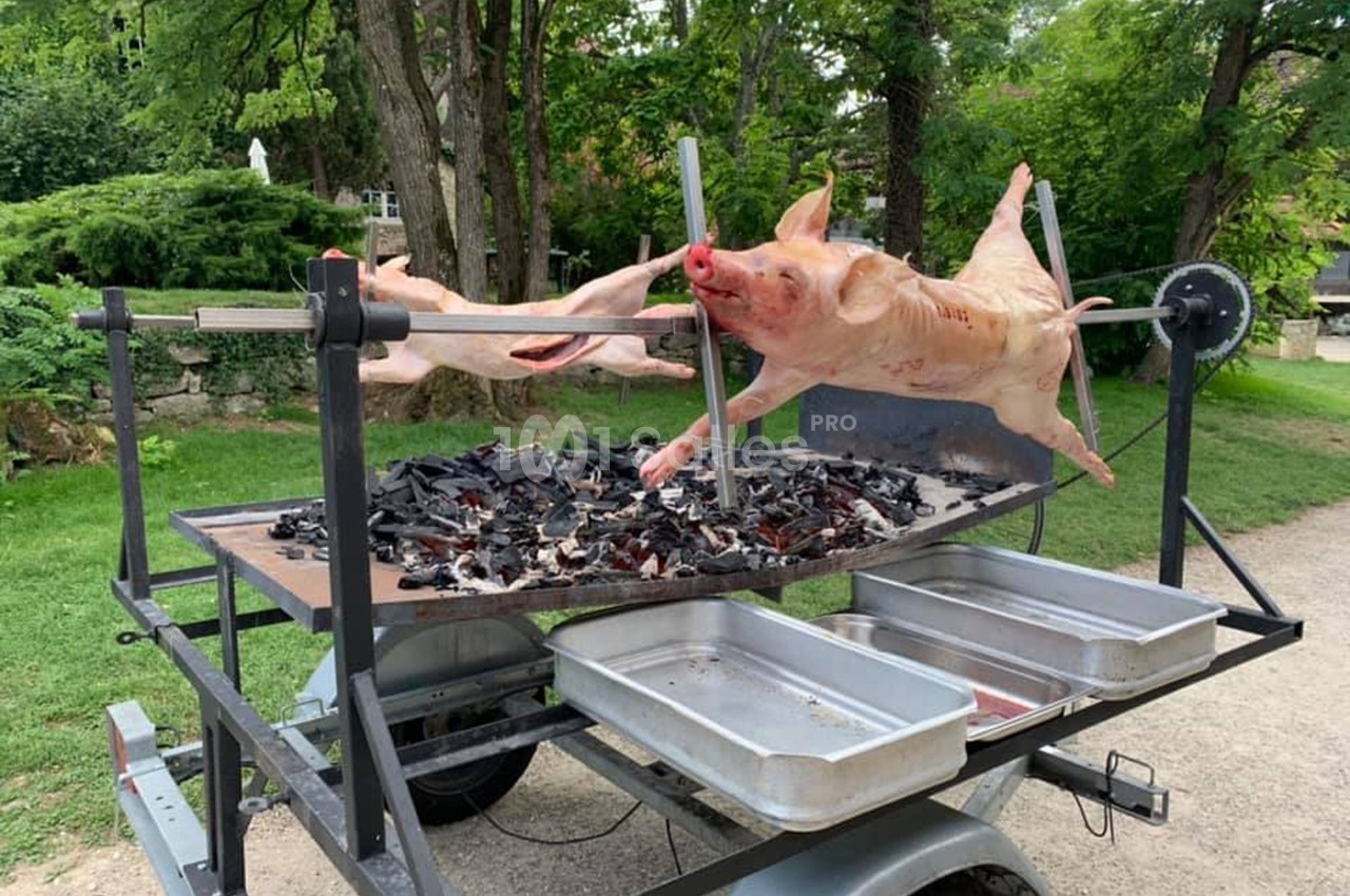 Cochon entier en cours de cuisson sur une broche rotative au-dessus de braises, dans un cadre extérieur verdoyant.