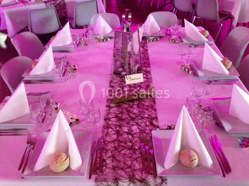 Table dressée pour un événement avec nappes blanches, serviettes pliées, couverts, verres et décoration centrale violette.