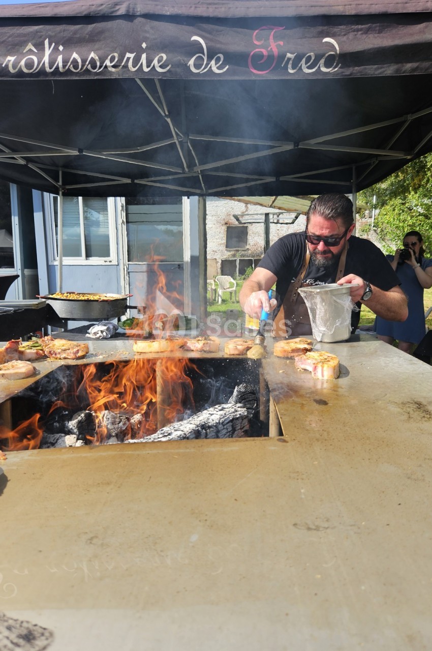 Un cuisinier prépare des plats sur un grill en plein air avec des flammes sous un auvent noir.