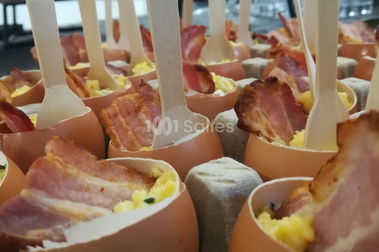 Œufs coquilles garnis d'œufs brouillés et de tranches de bacon, présentés avec des cuillères en bois.