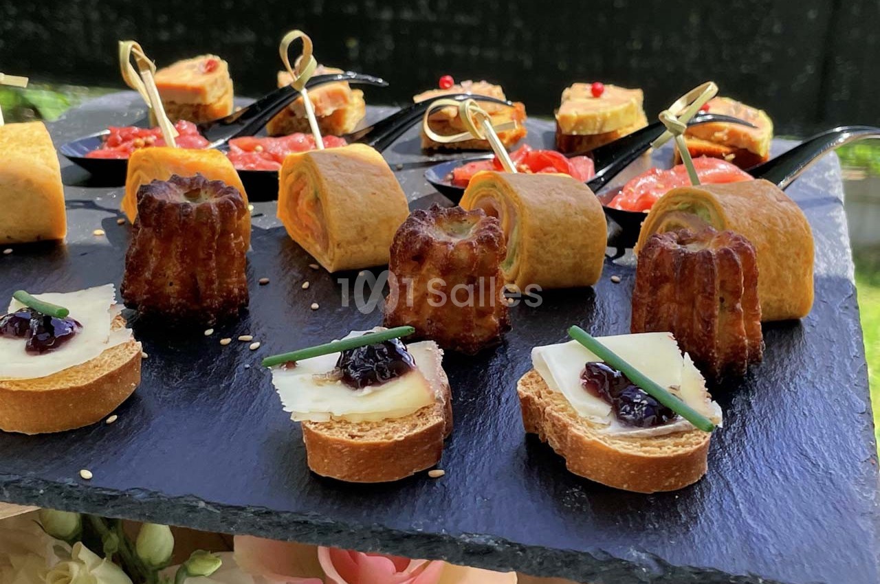 Plateau d'amuse-bouches variés comprenant cannelés, toasts garnis et roulés, présenté sur une ardoise noire.