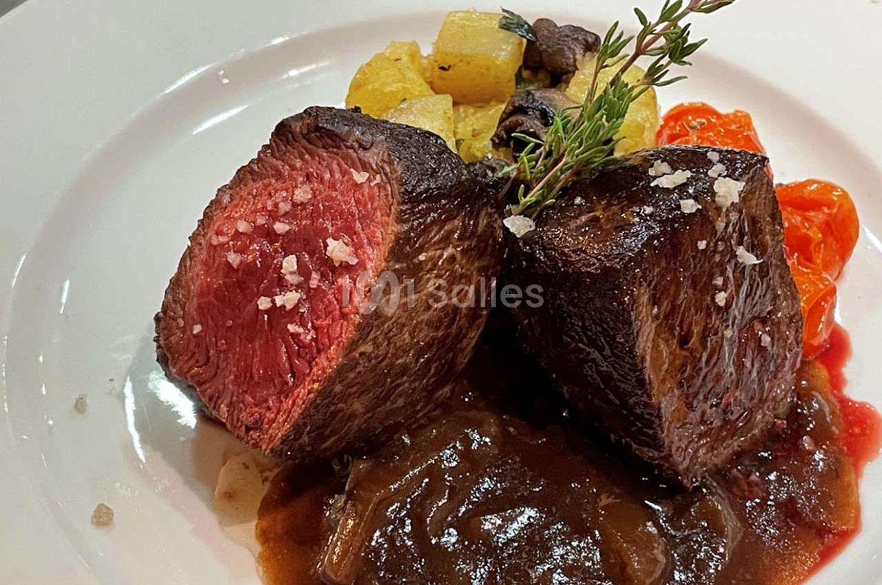 Deux morceaux de viande saignante servis avec sauce, légumes rôtis et herbes sur une assiette blanche.
