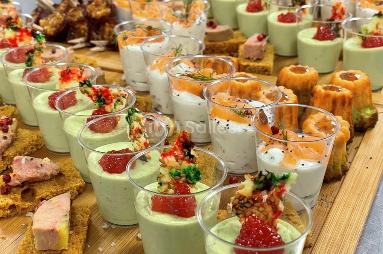 Plateau de verrines et amuse-bouches variés, incluant mousse verte, saumon, foie gras et cannelés, disposés sur un plateau…