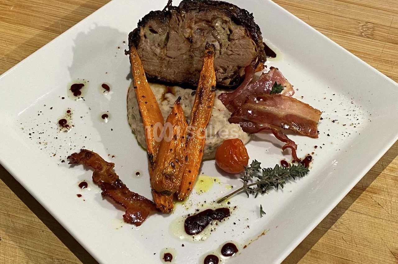 Assiette garnie d'un rôti de viande, purée, carottes rôties, bacon, tomate cerise et herbes sur une table en bois.