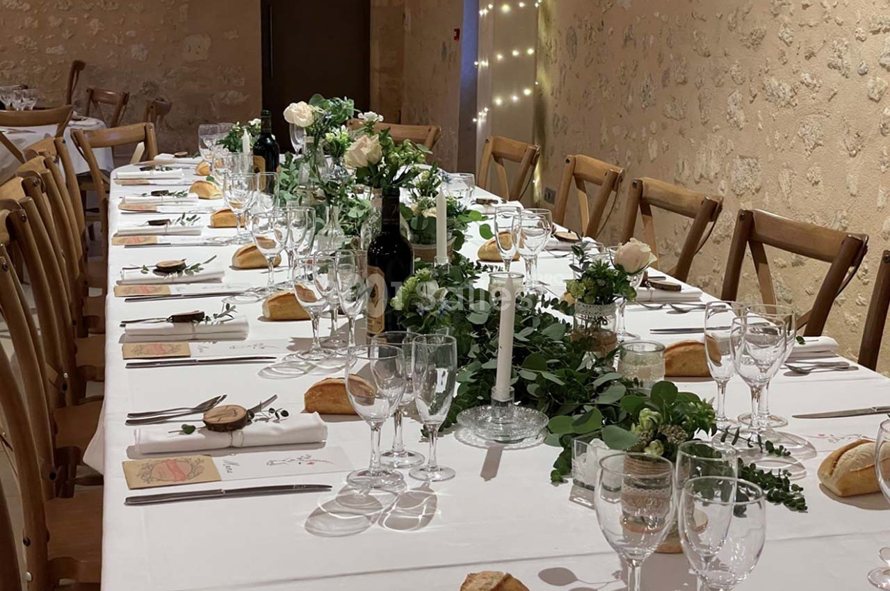 Table de réception élégamment dressée avec nappes blanches, bougies, fleurs et vaisselle, dans une salle en pierre.