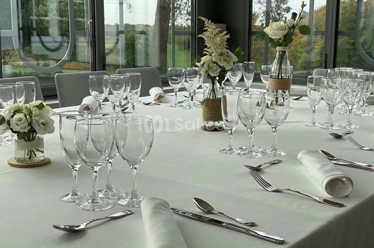 Table dressée avec nappes blanches, verres, couverts et décorations florales, dans une salle lumineuse avec vue sur l…
