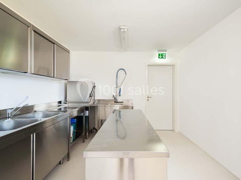 Cuisine professionnelle avec plans de travail en inox, évier, robinet flexible et porte de sortie au fond.