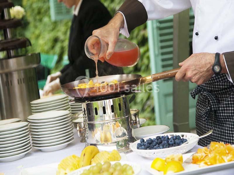 Un chef verse une sauce dans une poêle contenant des fruits, sur une table dressée avec des assiettes et des fruits frais.