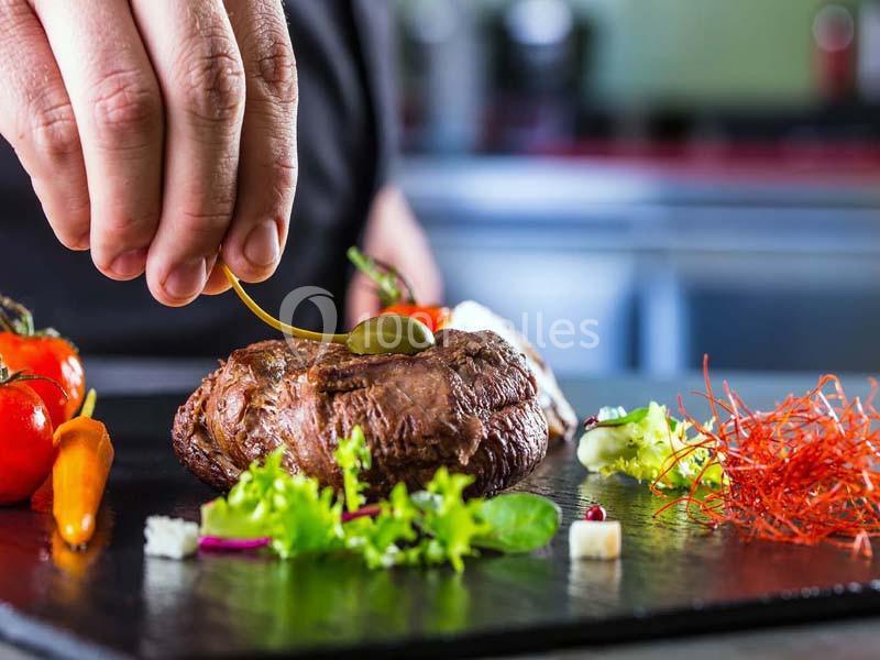 Un chef dépose une garniture sur un steak accompagné de légumes et de salade, présenté sur une assiette noire.
