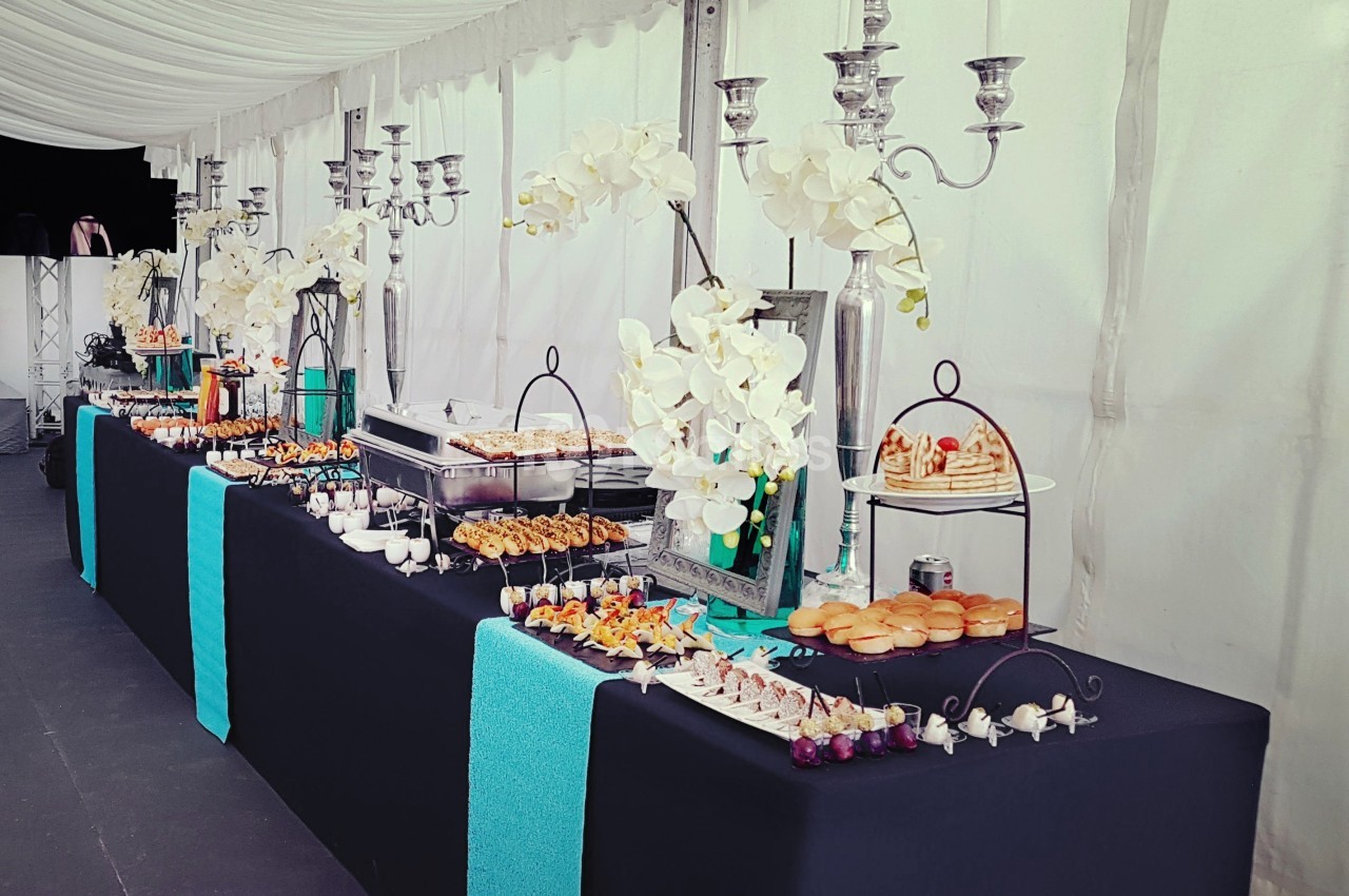 Buffet élégant avec nappes noires, décorations florales blanches et chandeliers, proposant divers plats et amuse-bouches.