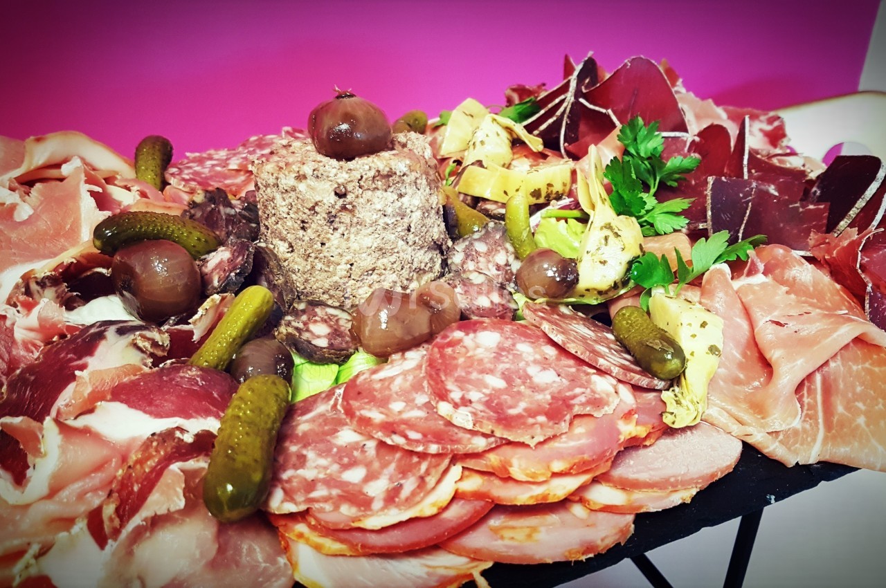 Plateau de charcuterie variée avec jambon, saucisson, cornichons, champignons et décorations sur fond rose.