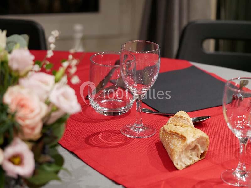 Table dressée avec nappe rouge, verres, couverts, serviette noire et morceau de baguette, fleurs en arrière-plan.