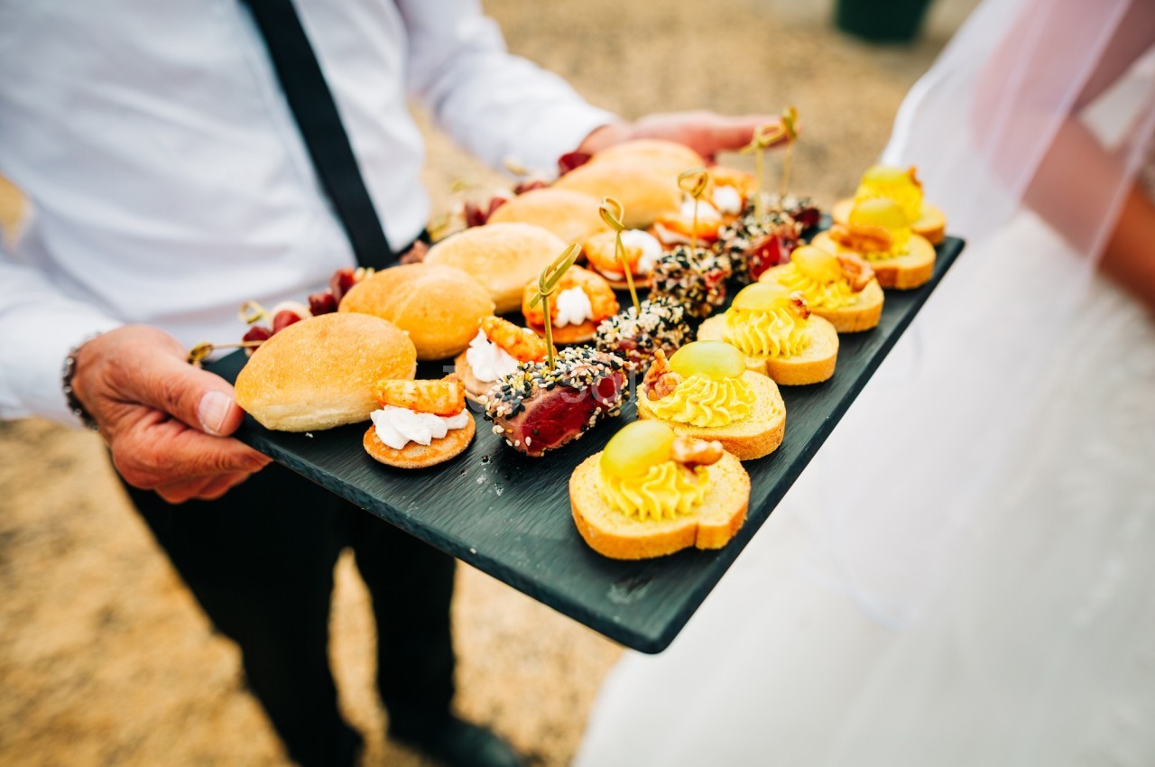 Plateau de bouchées apéritives variées, incluant mini-burgers, tartares et tartelettes, tenu par une personne en tenue…