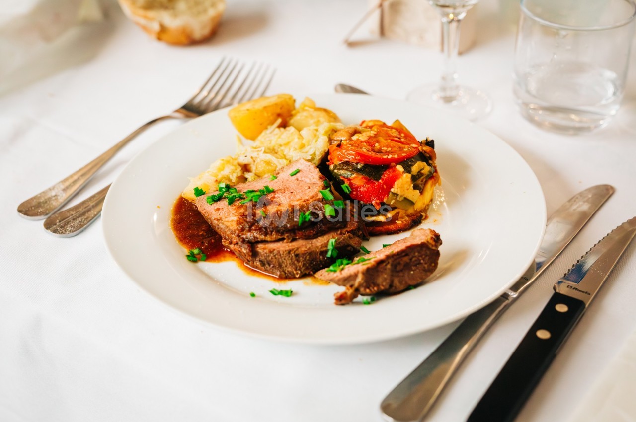 Assiette contenant une tranche de viande en sauce, légumes grillés, pommes de terre et riz, sur une table dressée.