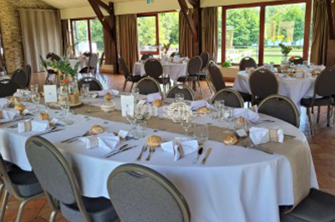 Salle de réception lumineuse avec tables dressées, nappes blanches, couverts et vue sur un jardin par de grandes baies…