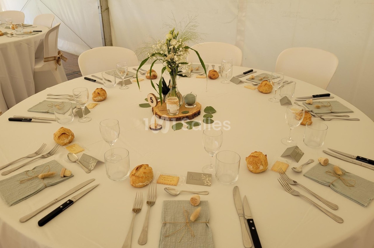 Table ronde dressée pour un repas, avec nappes blanches, couverts, serviettes, pains et décoration florale au centre.