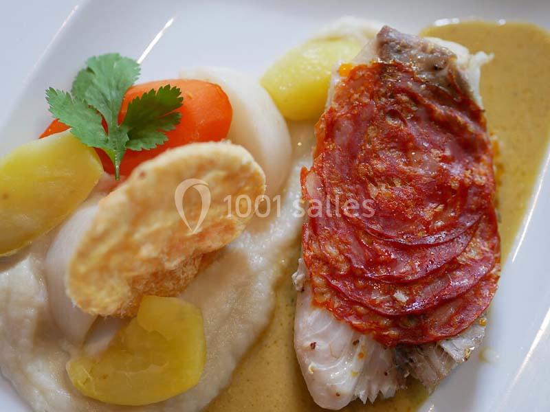 Plat composé de poisson garni de tranches de chorizo, purée, légumes et chips, présenté avec une sauce.