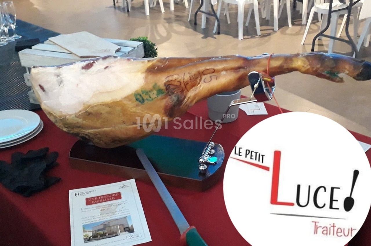 Un jambon entier sur support avec couteau, posé sur une table rouge, près de brochures et du logo ’Le Petit Luce Traiteur’.