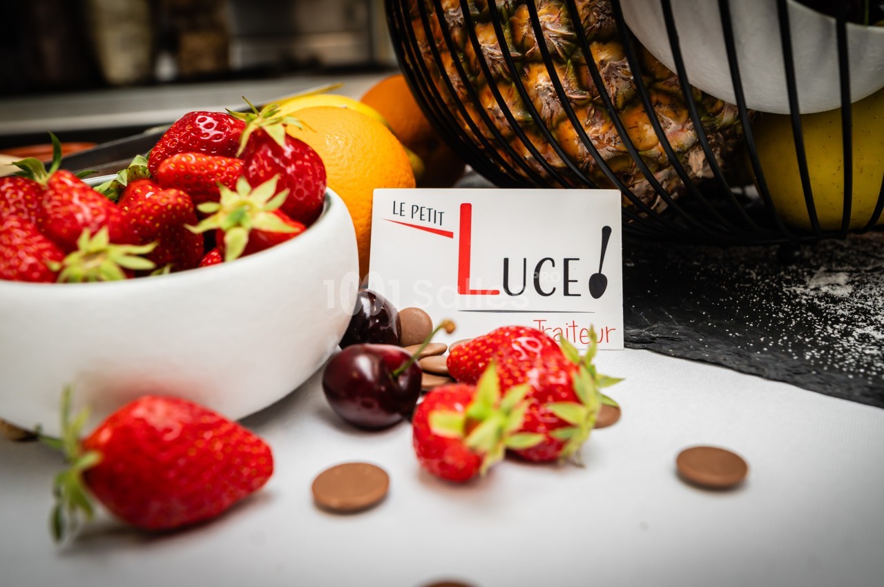 Bol de fraises fraîches avec cerises et chocolats, posé près d'un panier de fruits et une carte ’Le Petit Luce’.