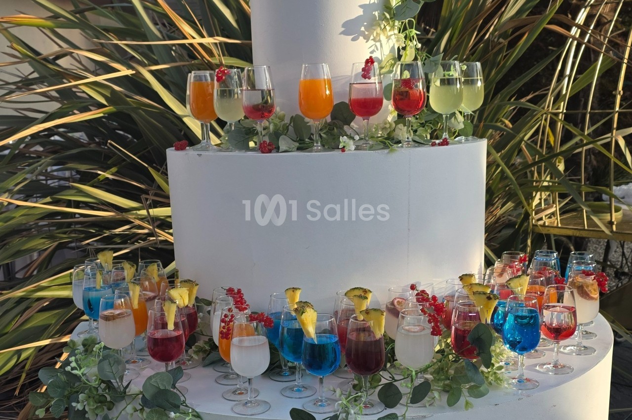 Présentoir de cocktails colorés décoré de feuillage et de fruits, disposé sur plusieurs niveaux en extérieur.