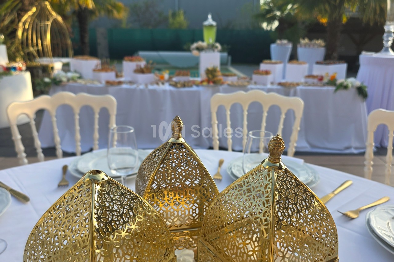 Table décorée avec des lanternes dorées ajourées, entourée de chaises blanches, avec un buffet en arrière-plan.