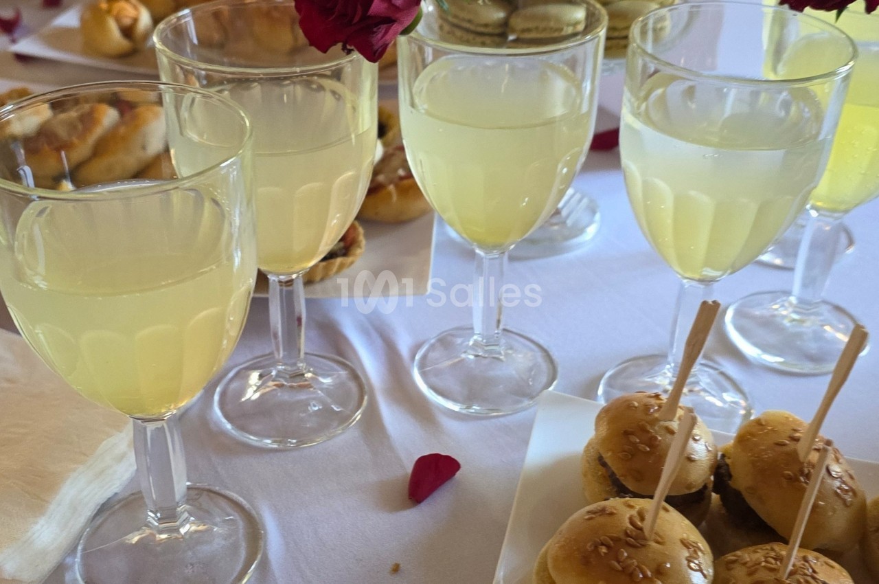 Verres de boisson jaune clair sur une table blanche, entourés de mini-burgers et de pétales de rose.