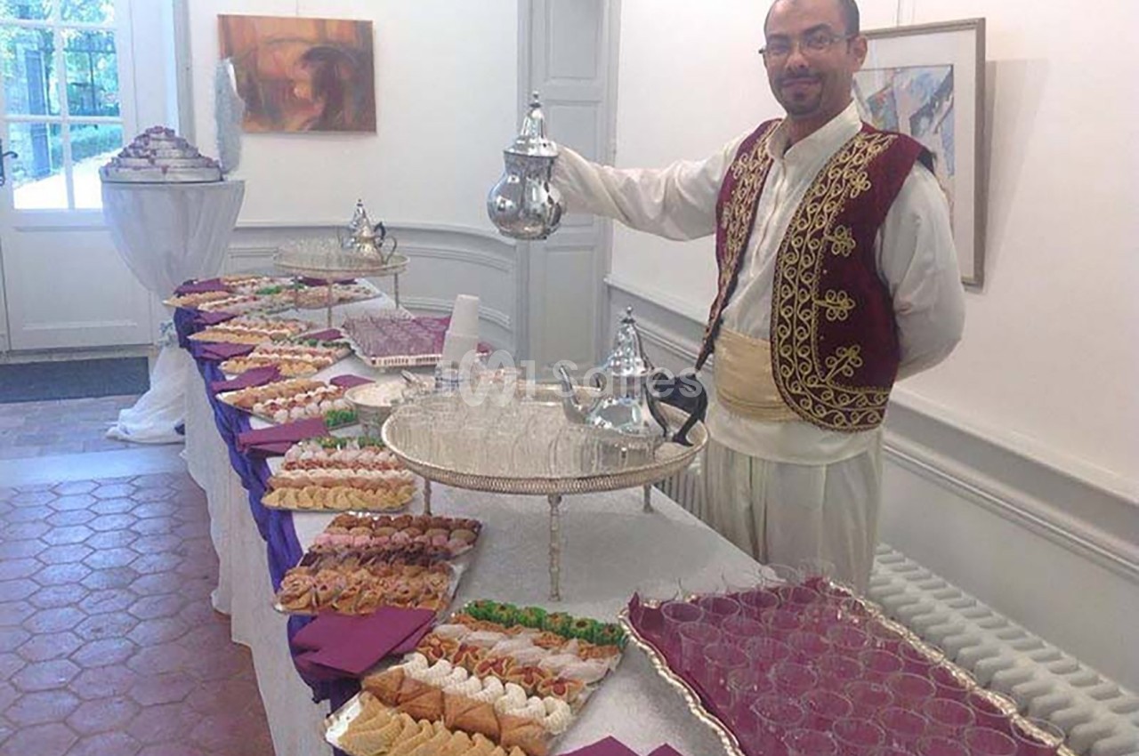 Un homme en tenue traditionnelle sert du thé près d'une table garnie de pâtisseries et de verres dans une salle lumineuse.