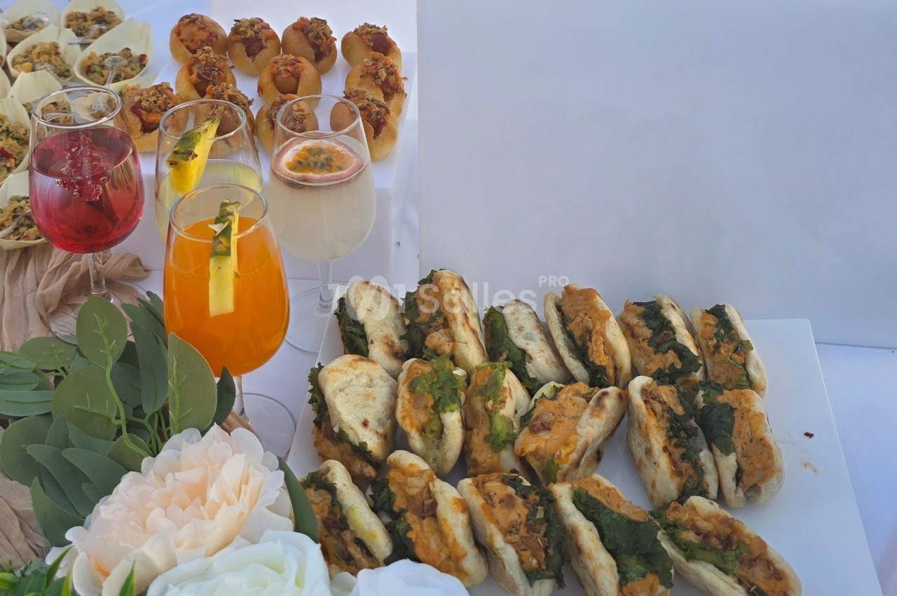 Plateau de mini-sandwichs garnis, accompagnés de cocktails colorés et de bouchées apéritives sur une table décorée.