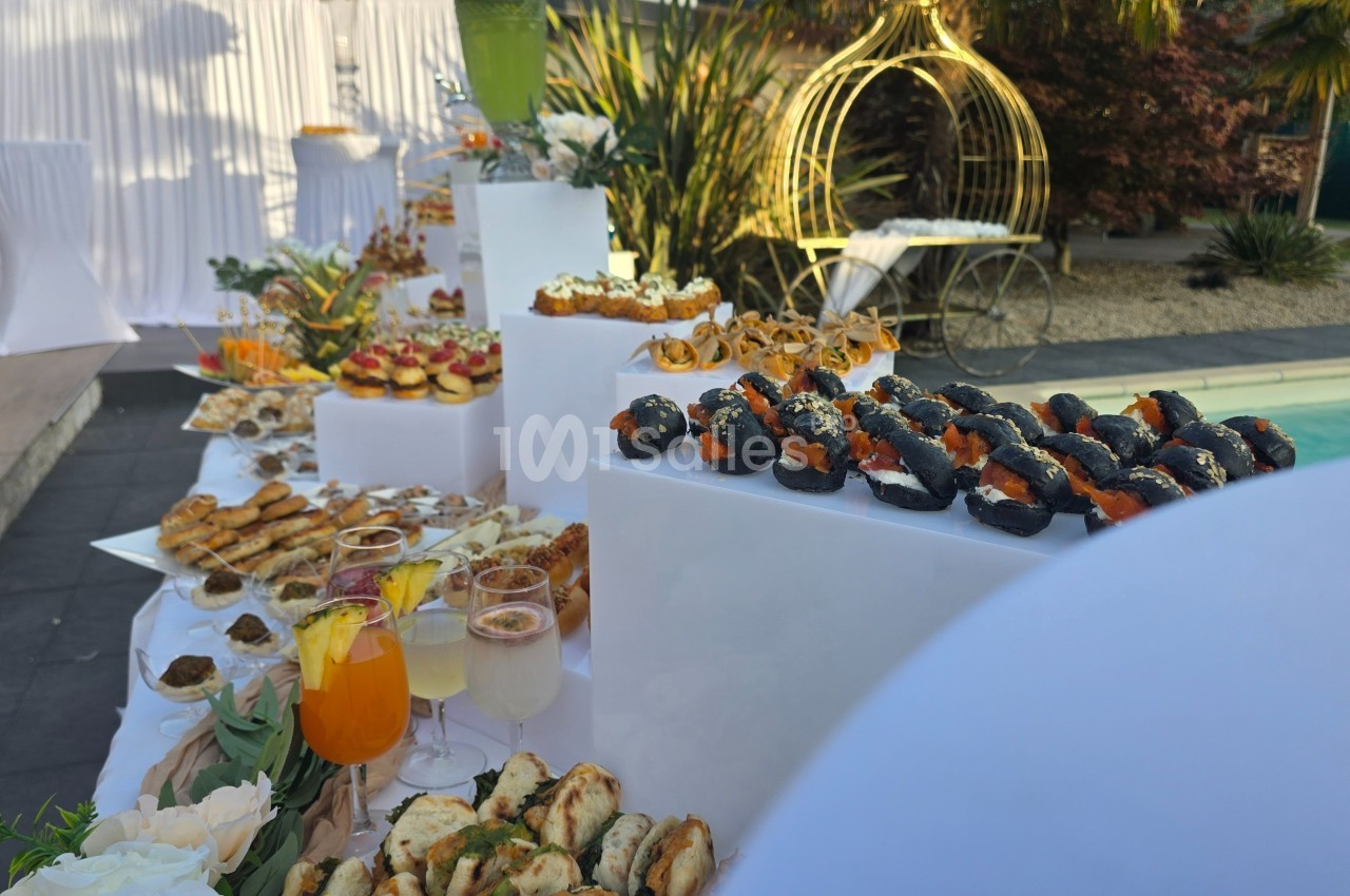 Buffet en extérieur avec divers amuse-bouches, verrines et boissons, disposés près d'une piscine et d'une décoration…