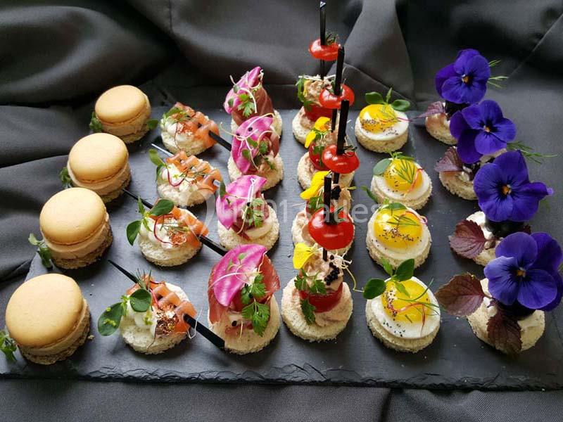 Plateau de canapés variés avec macarons salés, légumes, fleurs comestibles et décorations colorées sur ardoise noire.